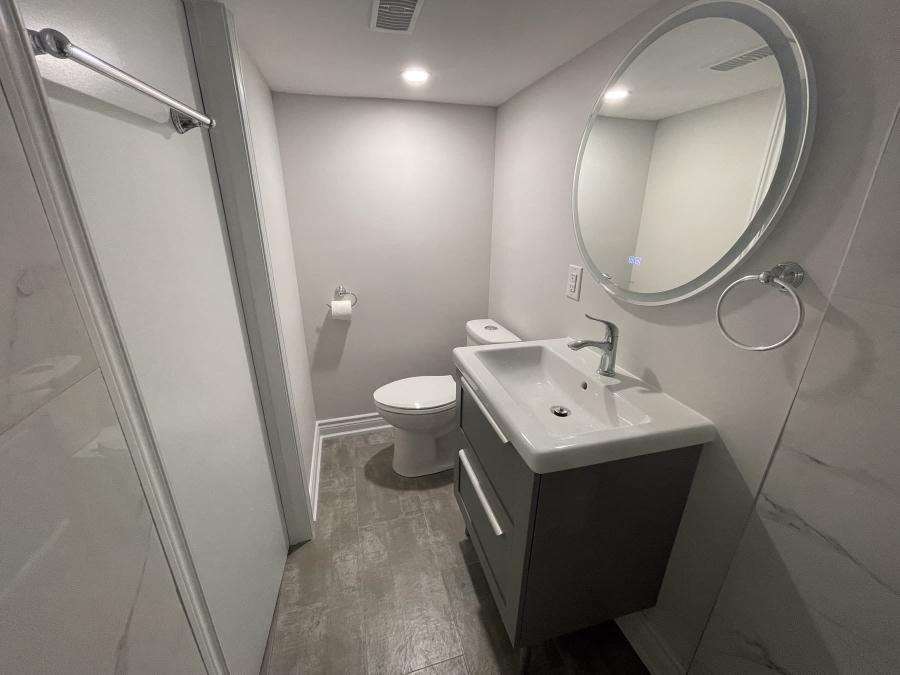 Finished bathroom view with grey vanity, round mirror, toilet, shower enclosure, and neutral wall finishes