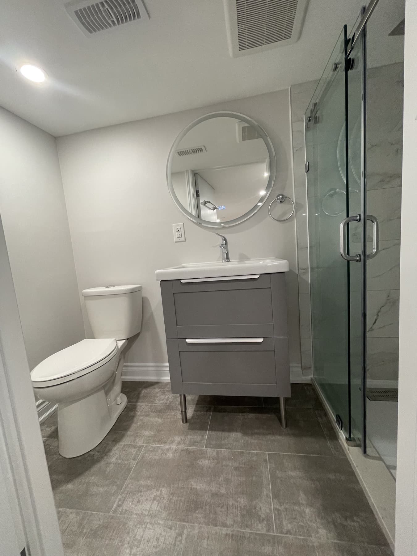 Finished compact bathroom with grey vanity, round mirror, toilet, glass shower door, and grey tile flooring